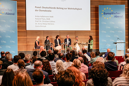 Eine Podiumsdiskussion mit sechs Personen auf einer Bühne vor einem vollen Publikum. Über dem Podium ist eine Leinwand mit dem Titel der Veranstaltung zu sehen: „Panel: Deutschlands Beitrag zur Wehrhaftigkeit der Demokratie“. Aufgeführt sind die Namen der Teilnehmenden: Britta Haßelmann MdB, Roland Theis MdB, Prof. Dr. Christian Calliess, Dr. Anna-Maija Mertens und Gosia Binczyk. Links und rechts auf der Bühne stehen Roll-Ups mit dem Logo „Europäischer Abend“.