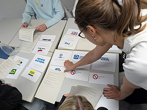 Mehrere Schüler:innen sitzen an Gruppentischen und ordnen Arbeitsblätter mit Logos europäischer Parteien wie „Renew Europe“, „Die Linke“, „ECR“ oder „Grüne/EFA“. Eine Lehrperson beugt sich über den Tisch und hilft bei der Zuordnung. Die Übung dient dem Verständnis der Fraktionen im Europäischen Parlament.