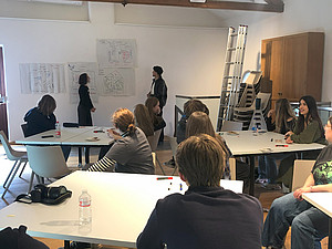 Das Bild zeigt eine Gruppe von Menschen in einem Raum, der wie ein Klassenzimmer oder ein Seminarraum eingerichtet ist. Die Teilnehmer sitzen an mehreren Tischen, die in einer U-Form angeordnet sind. Auf den Tischen liegen verschiedene Gegenstände wie Wasserflaschen, Marker und Notizblöcke. An der Wand hängen mehrere Flipcharts oder Poster mit Diagrammen und Notizen, die wahrscheinlich für eine Präsentation oder ein Brainstorming verwendet werden. Zwei Personen stehen vor den Flipcharts und scheinen eine Diskussion oder Präsentation zu leiten. Die Teilnehmer wirken aufmerksam und einige von ihnen sind in Gespräche vertieft oder machen sich Notizen. Im Hintergrund sind ein paar Stühle gestapelt und eine Leiter lehnt an der Wand. Es gibt auch eine Kamera auf einem der Tische, was darauf hindeuten könnte, dass die Veranstaltung dokumentiert wird. Der Raum ist hell und freundlich, mit großen Fenstern, die Tageslicht hereinlassen.